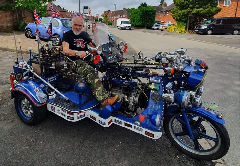 A man with a grey beard wearing a black t-shirt and combat trousers sitting on a motor-tricycle.