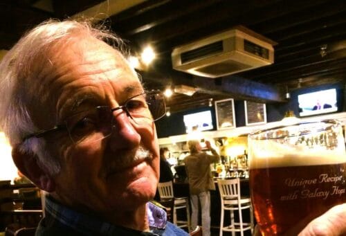 An older retired man holding a glass of beer smiling.