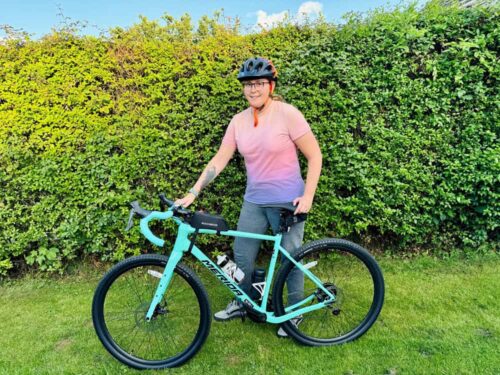 A young woman wearing cycling clothing, including a helmet, standing by a bicycle. She will be taking part in Birmingham Hospice's Ride the Reservoir fundraising event.